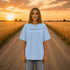 Woman wearing a white Comfort Colors t-shirt featuring a vintage prairie road and wheat field sunset graphic on the back while standing on a gravel road surrounded by golden wheat fields at sunset.