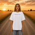 Woman wearing a white Comfort Colors t-shirt featuring a vintage prairie road and wheat field sunset graphic on the back while standing on a gravel road surrounded by golden wheat fields at sunset.