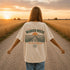 Woman wearing a white Comfort Colors t-shirt featuring a vintage prairie road and wheat field sunset graphic on the back while standing on a gravel road surrounded by golden wheat fields at sunset.