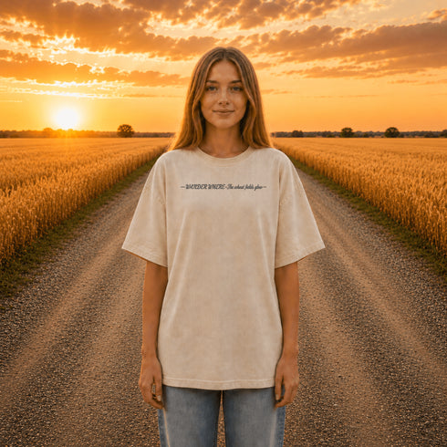 Woman wearing a white Comfort Colors t-shirt featuring a vintage prairie road and wheat field sunset graphic on the back while standing on a gravel road surrounded by golden wheat fields at sunset.