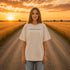 Woman wearing a white Comfort Colors t-shirt featuring a vintage prairie road and wheat field sunset graphic on the back while standing on a gravel road surrounded by golden wheat fields at sunset.