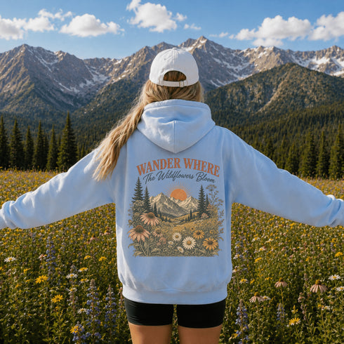 A person standing in a mountain meadow with arms outstretched, wearing a light blue hoodie with a large floral and mountain graphic on the back that reads “WANDER WHERE The Wildflowers Bloom,” plus a white cap, against a scenic alpine landscape.
