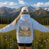 A person standing in a mountain meadow with arms outstretched, wearing a light blue hoodie with a large floral and mountain graphic on the back that reads “WANDER WHERE The Wildflowers Bloom,” plus a white cap, against a scenic alpine landscape.