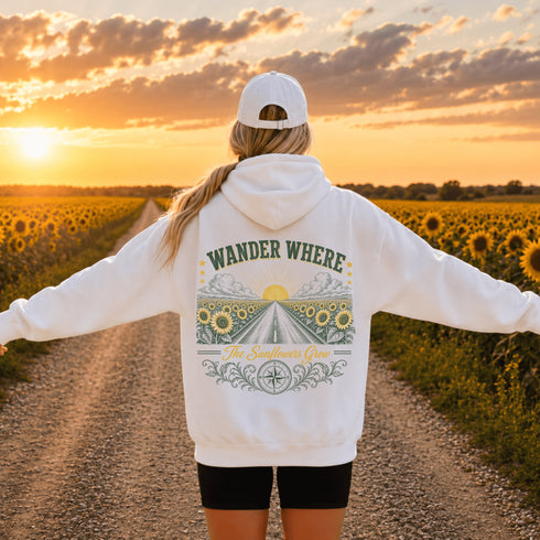 A woman stands on a dirt road between sunflower fields at sunset, viewed from behind. She wears a white hoodie with a large back graphic reading “WANDER WHERE The Sun Never Sets,” along with a white cap and black shorts.