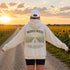 A person standing on a dirt path between sunflower fields at sunset, viewed from behind, wearing a cream hoodie with a large back graphic that reads “WANDER WHERE” above a road and sunflower illustration, plus a white cap.