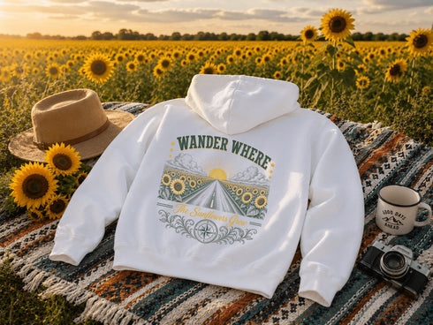 A white pullover hoodie laid flat on a woven blanket in a sunflower field at sunset, with a tan hat, mug, and camera placed around it. The hoodie has a large back graphic reading “WANDER WHERE” above a sunflower field scene and “The Sunflowers Grow” below.