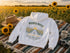 A white pullover hoodie laid flat on a woven blanket in a sunflower field at sunset, with a tan hat, mug, and camera placed around it. The hoodie has a large back graphic reading “WANDER WHERE” above a sunflower field scene and “The Sunflowers Grow” below.