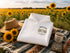 A folded white hoodie with a 'Wander Where Explore More' graphic is displayed on a colorful blanket in a sunflower field, styled with a straw hat, sunflowers, and a mug.