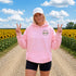 A woman stands on a dirt road between rows of sunflowers at sunset, wearing an oversized white hoodie with a small 'Wander where the sunflowers grow' chest logo and a white cap.