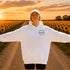 A woman stands on a dirt road between rows of sunflowers at sunset, wearing an oversized white hoodie with a small 'Wander where the sunflowers grow' chest logo and a white cap.