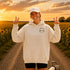 A woman stands on a dirt road between rows of sunflowers at sunset, wearing an oversized white hoodie with a small 'Wander where the sunflowers grow' chest logo and a white cap.