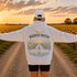 A person standing on a gravel path in a sunflower field at sunset, viewed from behind, wearing a light gray hoodie with a large graphic print that reads “WANDER WHERE” above a road and sunflower illustration, along with a white cap and black shorts.