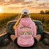 A woman sits in the bed of a rustic green pickup truck facing away from the camera, wearing a pink hoodie with a sunflower field and road graphic on the back, a white baseball cap, and black shorts. The background shows a golden sunset over a field of sunflowers.