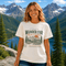 Woman wearing Wander Far Take the Scenic Route mountain hiking graphic t-shirt featuring vintage mountain landscape artwork with pine trees and sunset background.