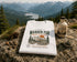 White t-shirt with nature-themed design on a rock with scenic mountains and lake in the background