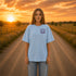 Woman wearing a Comfort Colors 1717 t-shirt featuring a Wander Where the Wildflowers Grow prairie sunset and wildflower field back graphic while standing on a gravel road at sunset.
