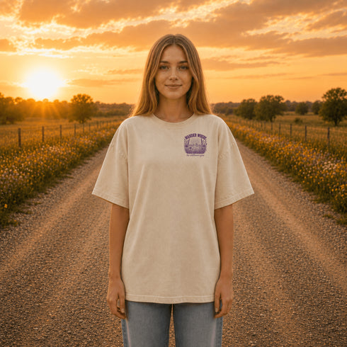 Woman wearing a Comfort Colors 1717 t-shirt featuring a Wander Where the Wildflowers Grow prairie sunset and wildflower field back graphic while standing on a gravel road at sunset.