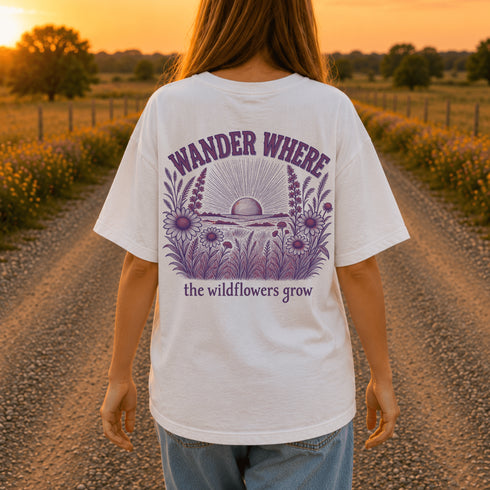 Woman wearing a Comfort Colors 1717 t-shirt featuring a Wander Where the Wildflowers Grow prairie sunset and wildflower field back graphic while standing on a gravel road at sunset.