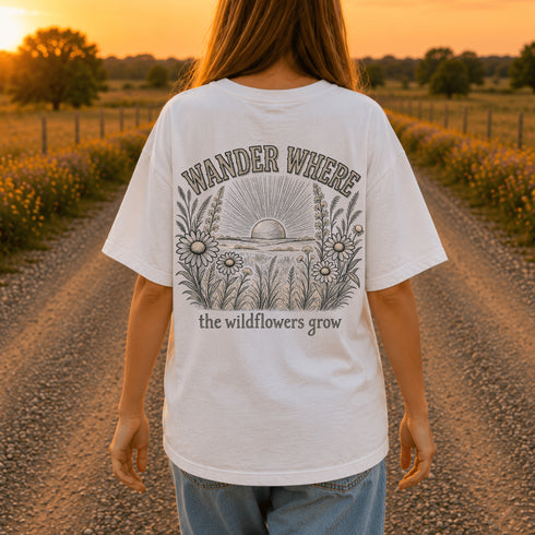 Woman wearing a Comfort Colors 1717 t-shirt featuring a Wander Where the Wildflowers Grow prairie sunset and wildflower field back graphic while standing on a gravel road at sunset.