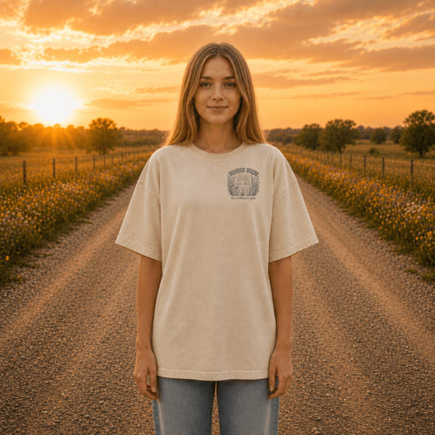 Woman wearing a Comfort Colors 1717 t-shirt featuring a Wander Where the Wildflowers Grow prairie sunset and wildflower field back graphic while standing on a gravel road at sunset.