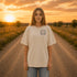 Woman wearing a Comfort Colors 1717 t-shirt featuring a Wander Where the Wildflowers Grow prairie sunset and wildflower field back graphic while standing on a gravel road at sunset.