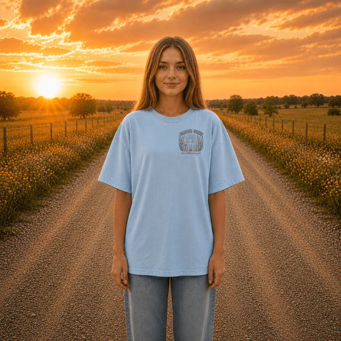 Woman wearing a Comfort Colors 1717 t-shirt featuring a Wander Where the Wildflowers Grow prairie sunset and wildflower field back graphic while standing on a gravel road at sunset.