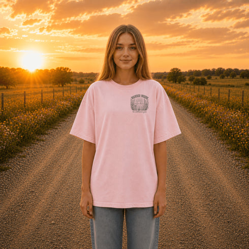 Woman wearing a Comfort Colors 1717 t-shirt featuring a Wander Where the Wildflowers Grow prairie sunset and wildflower field back graphic while standing on a gravel road at sunset.