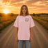 Woman wearing a Comfort Colors 1717 t-shirt featuring a Wander Where the Wildflowers Grow prairie sunset and wildflower field back graphic while standing on a gravel road at sunset.