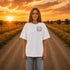 Woman wearing a Comfort Colors 1717 t-shirt featuring a Wander Where the Wildflowers Grow prairie sunset and wildflower field back graphic while standing on a gravel road at sunset.