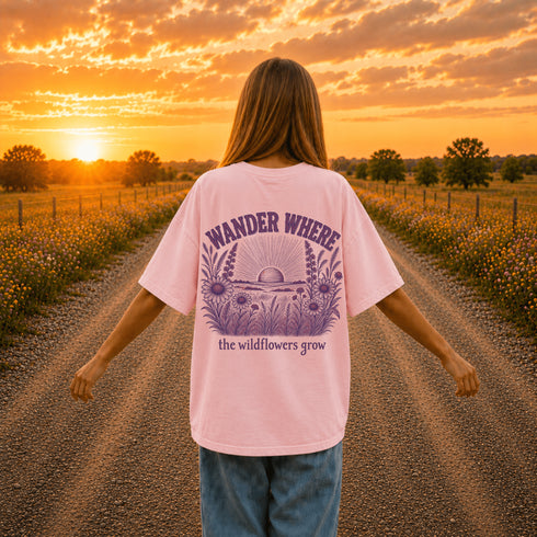 Woman wearing a Comfort Colors 1717 t-shirt featuring a Wander Where the Wildflowers Grow prairie sunset and wildflower field back graphic while standing on a gravel road at sunset.