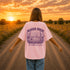 Woman wearing a Comfort Colors 1717 t-shirt featuring a Wander Where the Wildflowers Grow prairie sunset and wildflower field back graphic while standing on a gravel road at sunset.