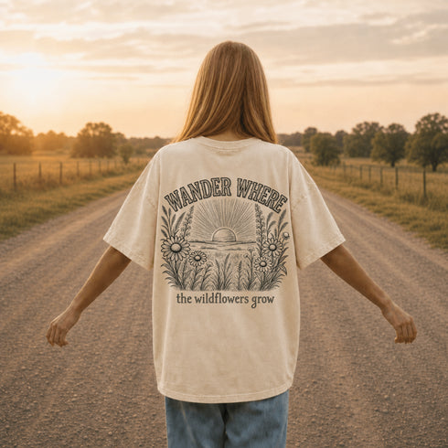 Woman wearing a Comfort Colors 1717 t-shirt featuring a Wander Where the Wildflowers Grow prairie sunset and wildflower field back graphic while standing on a gravel road at sunset.