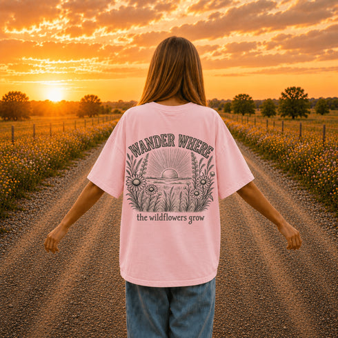 Woman wearing a Comfort Colors 1717 t-shirt featuring a Wander Where the Wildflowers Grow prairie sunset and wildflower field back graphic while standing on a gravel road at sunset.