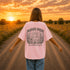 Woman wearing a Comfort Colors 1717 t-shirt featuring a Wander Where the Wildflowers Grow prairie sunset and wildflower field back graphic while standing on a gravel road at sunset.