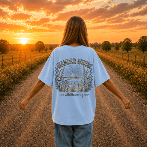 Woman wearing a Comfort Colors 1717 t-shirt featuring a Wander Where the Wildflowers Grow prairie sunset and wildflower field back graphic while standing on a gravel road at sunset.