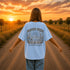 Woman wearing a Comfort Colors 1717 t-shirt featuring a Wander Where the Wildflowers Grow prairie sunset and wildflower field back graphic while standing on a gravel road at sunset.