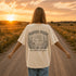 Woman wearing a Comfort Colors 1717 t-shirt featuring a Wander Where the Wildflowers Grow prairie sunset and wildflower field back graphic while standing on a gravel road at sunset.
