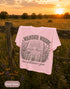 Pink t-shirt with 'Wander where the wildflowers grow' design hanging on a wooden fence in a field at sunset.
