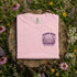 Pink sweatshirt with a purple graphic and text on a wooden stump surrounded by wildflowers