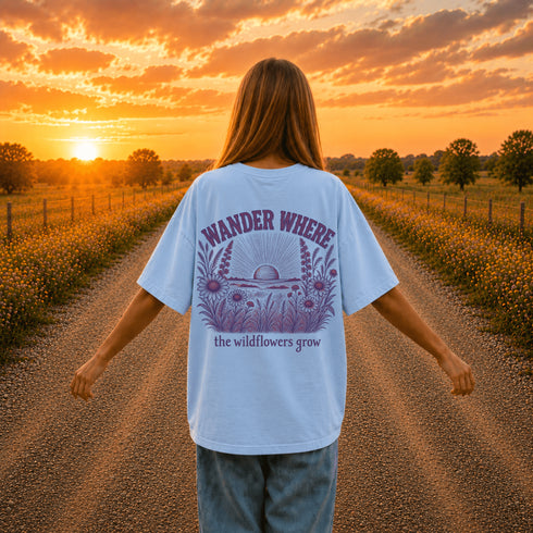 Woman wearing a Comfort Colors 1717 t-shirt featuring a Wander Where the Wildflowers Grow prairie sunset and wildflower field back graphic while standing on a gravel road at sunset.