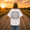 Woman wearing a Comfort Colors 1717 t-shirt featuring a Wander Where the Wildflowers Grow prairie sunset and wildflower field back graphic while standing on a gravel road at sunset.