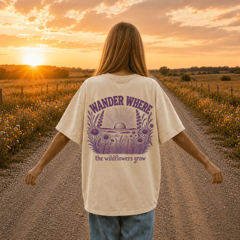 Woman wearing a Comfort Colors 1717 t-shirt featuring a Wander Where the Wildflowers Grow prairie sunset and wildflower field back graphic while standing on a gravel road at sunset.