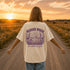 Woman wearing a Comfort Colors 1717 t-shirt featuring a Wander Where the Wildflowers Grow prairie sunset and wildflower field back graphic while standing on a gravel road at sunset.