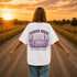 Woman wearing a Comfort Colors 1717 t-shirt featuring a Wander Where the Wildflowers Grow prairie sunset and wildflower field back graphic while standing on a gravel road at sunset.