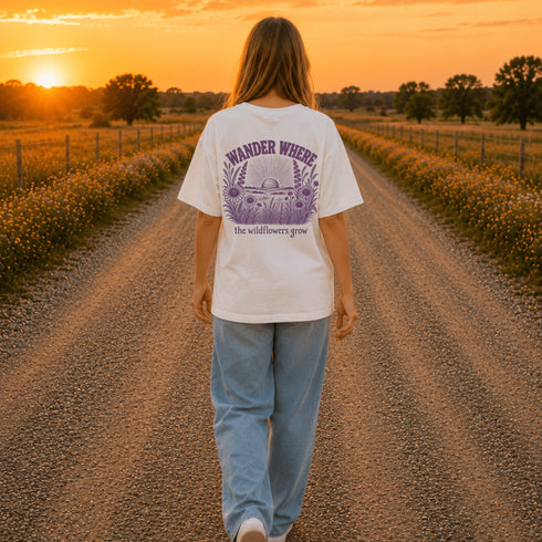 Woman wearing a Comfort Colors 1717 t-shirt featuring a Wander Where the Wildflowers Grow prairie sunset and wildflower field back graphic while walking on a gravel road at sunset.