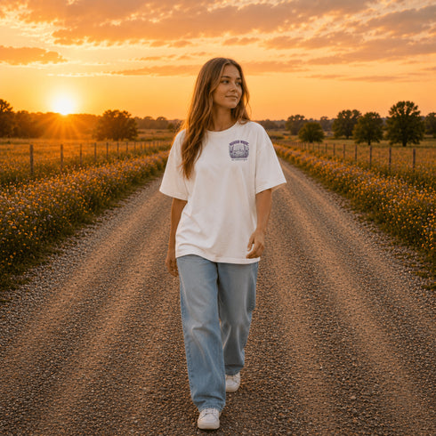 Woman wearing a Comfort Colors 1717 t-shirt featuring a Wander Where the Wildflowers Grow prairie sunset and wildflower field back graphic while standing on a gravel road at sunset.