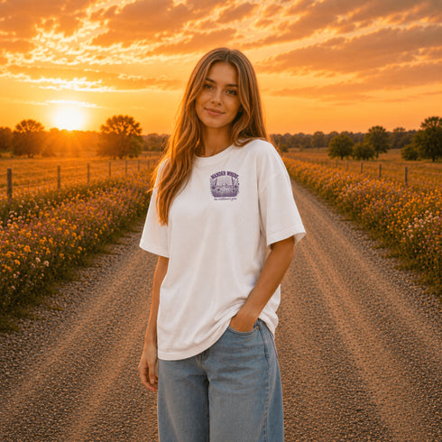 Woman wearing a Comfort Colors 1717 t-shirt featuring a Wander Where the Wildflowers Grow prairie sunset and wildflower field back graphic while standing on a gravel road at sunset.