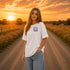 Woman wearing a Comfort Colors 1717 t-shirt featuring a Wander Where the Wildflowers Grow prairie sunset and wildflower field back graphic while standing on a gravel road at sunset.