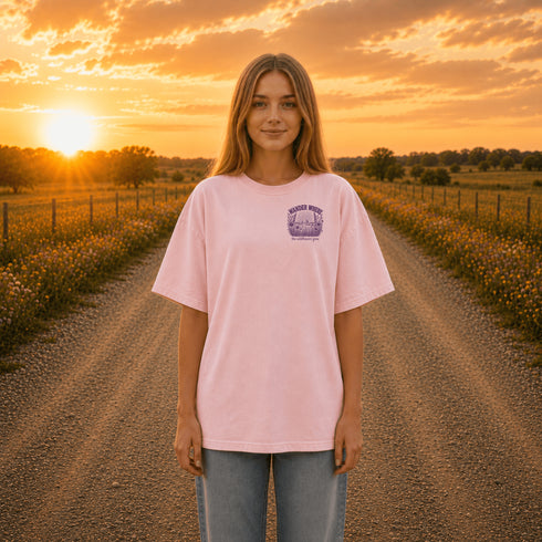 Person wearing a pink t-shirt with a logo standing on a dirt road during sunset.
