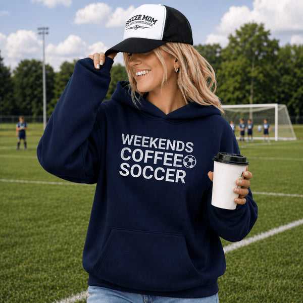Woman wearing a navy hoodie with 'Weekends Coffee Soccer' text, holding a coffee cup on a soccer field.